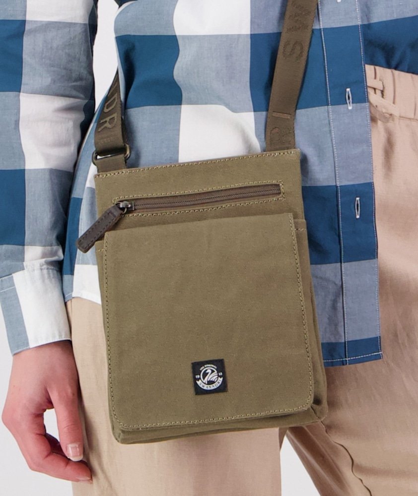 An olive green canvas crossbody bag with a front zip pocket and a small black logo patch, shown worn across the chest of a person wearing a blue and white plaid shirt and beige pants.