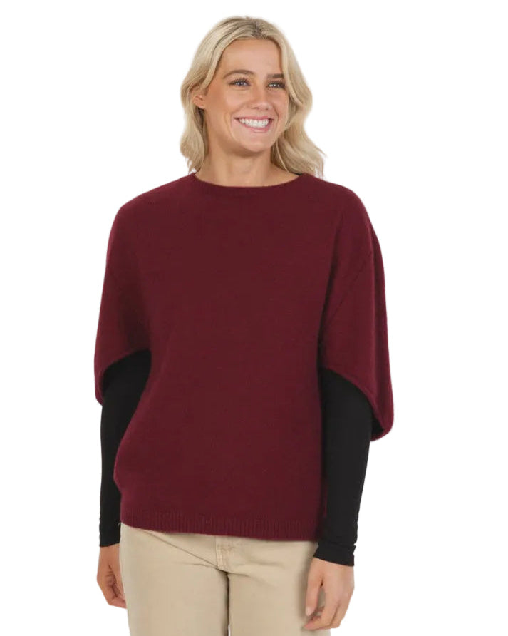 Woman wearing a burgundy poncho over a black top with beige pants on a white background