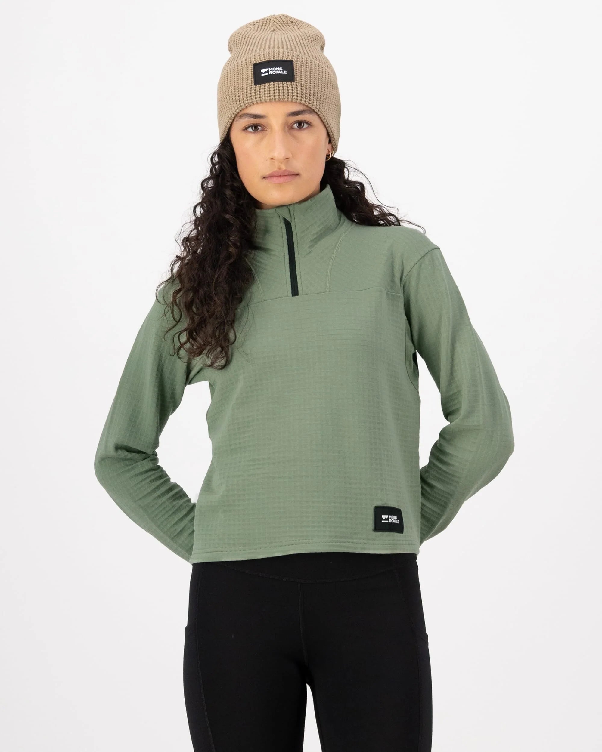 Woman wearing a green pullover, black leggings, and a beige beanie with a logo on a white background